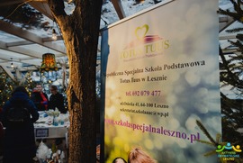 Zdjęcie przedstawia banner Niepublicznej Specjalnej Szkoły Podstawowej Totus Tuus w Lesznie. W tle są kobiety sprzedające wyroby (photo)
