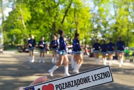 Zdjęcie przedstawia na pierwszym planie tabliczkę z napisem I love pozarządowe Leszno. W tle widać rozmyte sylwetki dziewczynek w strojach mażoretek. (photo)