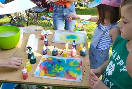 Zdjęcie przedstawia stoisko animacyjne dla dzieci. Znajdują się na ni farby i deska do łączenia kolorów. Dookoła stoją dzieci wpatrujące się w kolory. (photo)