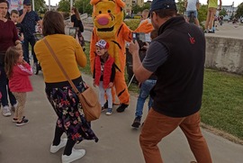 Zdjęcie przedstawia maskotkę Tygryska, dziecko, które pozuje z nim do zdjęcia, dorosłych robiących zdjęcia oraz kilku uczestników Miejskiego Dnia Dziecka.  (photo)