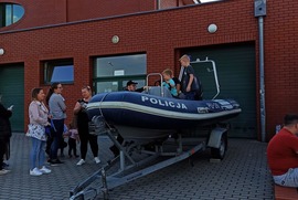 Zdjęcie przedstawia ponton policyjny, dzieci w nim siedzące, policjanta, który tłumaczy dzieciom co znajduje się z pontonie oraz kilku uczestników Miejskiego Dnia Dziecka stojących obok. (photo)